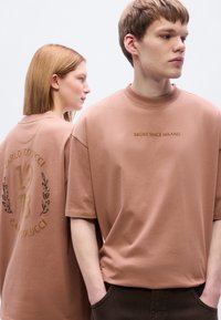 Καφέ oversized μπλουζάκια με ριμπ λαιμόκοψη. Μπροστινό κείμενο: «BROKE SINCE MILANO.» Πλάτη με διακοσμητική κεντητή επιγραφή: «CARLO COLUCCI 1973.»