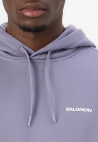 Hoodie violet clair en tissu doux, doté d'une poche avant, d'une capuche avec cordon de serrage et du logo blanc "SALOMON" sur la poitrine.