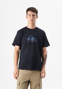 Jaunas vyras stovi pasitikintis savimi, vilkėdamas tamsiai mėlyną Quiksilver marškinėlį su logotipu, derinant su kuprinėmis kišenėmis kelnais, prieš baltą fono.