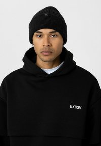 Hoodie noir avec un design superposé et une poche avant ; présente un logo graphique blanc. Un bonnet en tricot côtelé noir complète la tenue.