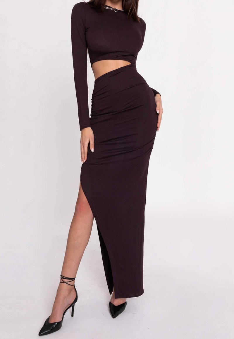 Femme portant un crop top noir à manches longues et une jupe taille haute assortie avec une fente haute sur la cuisse, associée à des talons hauts noirs pointus.