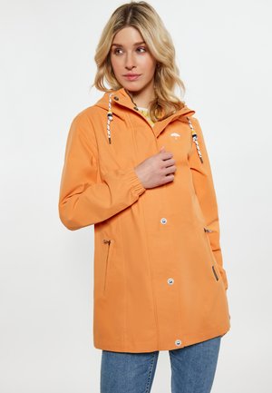 Blondynka w pomarańczowym kurtce przeciwdeszczowej z kapturem i kieszeniami na suwak oraz w niebieskich dżinsach, patrzy na bok na jednolitym białym tle.