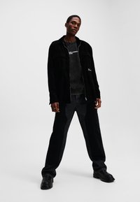 Sort fløjl zip-up jakke med to frontlommer, lagd over en mørk grafisk t-shirt. Sorte denimbukser og kraftige sorte sko.