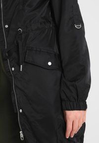 Czarna nylonowa kurtka z zapięciem na zamek, z regulacją w talii i elastycznymi mankietami. Zawiera kieszeń z zapięciem na nap. 