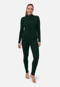 Dunkelgrünes langärmliges Oberteil und passende Leggings in figurbetontem Design. Weiche Textur mit kontrastierenden Steppnähten. Modell steht barfuß.