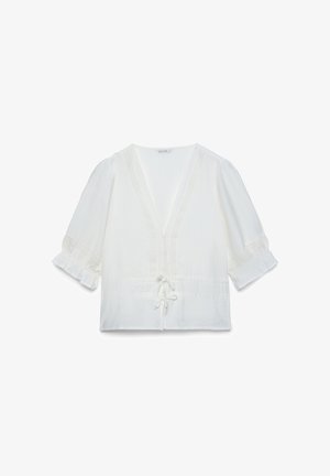 Blusa bianca con scollo a V, maniche a sbuffo 3/4, cuciture decorative lungo lo scollo e le maniche, e fiocco frontale in vita.