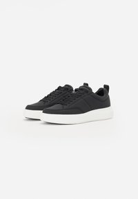 Des baskets noires avec une tige synthétique lisse, une semelle en caoutchouc blanche, une forme de bout arrondie, un système de laçage et une languette au talon.