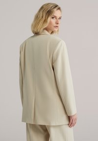 Béžové sako s hladkou textúrou, jedným zadným rozparkom, štruktúrovanými ramenami a dlhými rukávmi, doplnené o dizajn s otvoreným golierom.