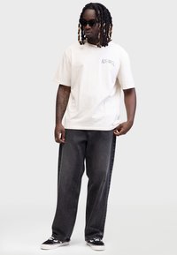 Hellbeige Baumwoll-T-Shirt mit kurzen Ärmeln, versehen mit einem bunten Graphic auf der linken Brust. Kombiniert mit dunkelgrauen weiten Jeans und schwarzen Sneakers.