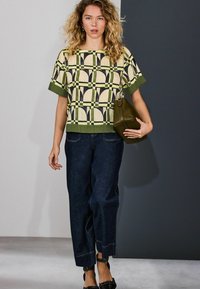 Lühikeste varrukatega top, millel on kollase ja rohelise geomeetriline muster, kombineeritud tumeliste laia säärega teksadega ja tumerohelise käekotiga.