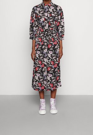 Frau trägt ein schwarzes Midikleid mit rotem, weißem und lila Blumenmuster, langen Ärmeln und lila High-Top-Sneakern, steht vor einem schlichten Hintergrund.