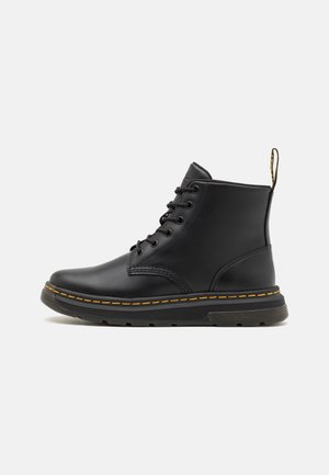 Bottes cheville en cuir noir avec bout arrondi, design à lacets, couture jaune le long de la semelle et une languette en tissu à l'arrière.