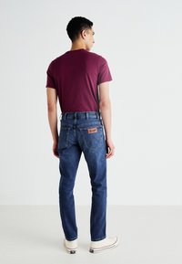 Vīrietis ar muguru pret skatītāju, tērpies bordo krāsas T-kreklā, zilās Wrangler džinsās un baltos augstajos sporta apavos, pret tukšu baltu fona.