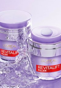 Crèmes en pots L'Oréal Paris Revitalift Filler, en verre transparent, avec des accents violets et des couvercles argentés, avec une goutte de crème sur le couvercle, sur fond texturé.