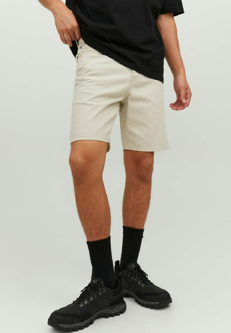 Pantaloni corti beige con taglio dritto e orlo, abbinati a una t-shirt nera, calzini neri e sneakers nere. Tessuto morbido, design casual.