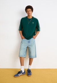 Grøn kortærmet t-shirt med hvide tre-stribe detaljer, lyseblå denim shorts og marineblå sneakers med hvide accenter.