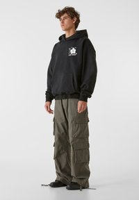 Fekete kapucnis pulóver elülső zsebel és fehér felhő grafikával, olajzöld cargo nadrággal párosítva, amely több zsebbel és húzózsinórral rendelkezik.