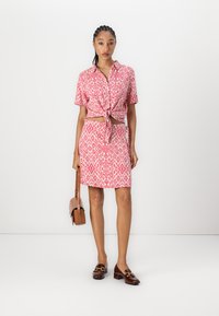 Robe en coton mélangé rouge et blanc avec un motif géométrique, manches courtes, devant noué et ceinture assortie. Mocassins marron et petit sac.