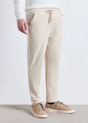 Uomo che indossa pantaloni della tuta beige con coulisse in vita, sneakers beige e maglione bianco a costine, in piedi su pavimento chiaro.