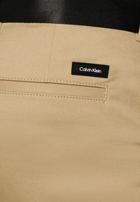 Pantaloni beige con una texture liscia, caratterizzati da un'etichetta nera di Calvin Klein sulla tasca posteriore e dettagli cuciti lungo le cuciture.