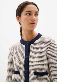 Knitted cardigan in cream and navy with a textured pattern, a round neck, and two front pockets. Features four blue buttons.