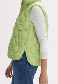 Puchowa bezrękawnik w jasnym zielonym kolorze z pikowanym wzorem, z boczną kieszenią i wysokim kołnierzem. Noszony na białej koszuli z długim rękawem.