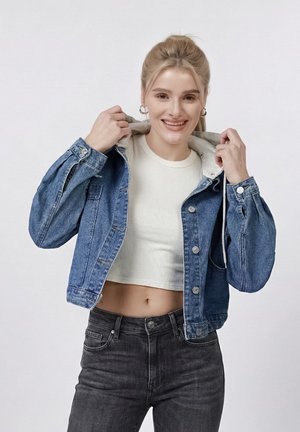 Jeune femme souriante ajustant le col de sa veste en jean bleue portée sur un haut blanc court et un jean noir.