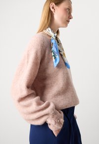 Zachte roze gebreide trui met losse, puffy mouwen en knoopaccenten. Gecombineerd met een zijden sjaal met bloemen in blauw en wit. Marineblauwe broek.