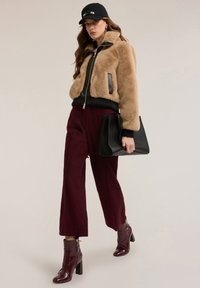 Kobieta w beżowej kurtce z sztucznego futra, bordo spodniach, dopasowanych botkach, czapce i trzymająca dużą czarną torebkę na jednolitym tle.