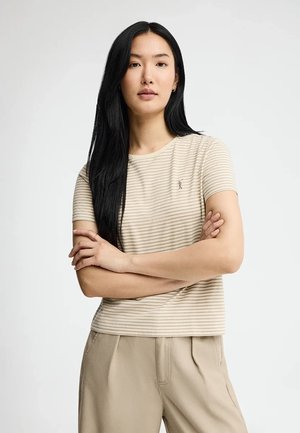 Jeune femme aux longs cheveux noirs portant une chemise à manches courtes beige rayée et un pantalon beige clair, bras croisés, expression neutre.