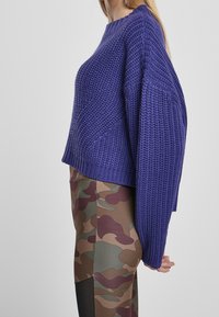 Överdimensionerad lila stickad tröja med ett texturerat mönster, rund halsringning och förlängda ärmar, tillsammans med byxor i camouflage-mönster.