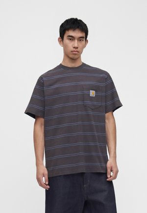 Jeune homme portant un t-shirt gris foncé à manches courtes avec de fines rayures horizontales bleues et une poche poitrine avec un patch logo Carhartt.