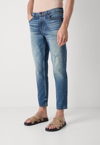 Jeans azul claro con un acabado deslavado, con un corte slim y detalles desgastados, combinados con sandalias de tiras marrones sobre un fondo neutro.