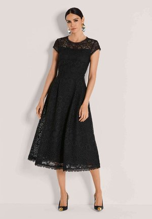Schwarzes Spitzenkleid mit kurzen Ärmeln, floralem Ausschnitt-Design, ausgestelltem Rock und gewelltem Saum. Tragen mit schwarzen Heels mit goldenen Akzenten.