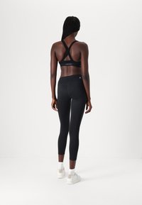 Schwarzer Sport-BH mit überkreuzten Trägern, kombiniert mit hoch taillierten schwarzen Leggings. Das Outfit zeichnet sich durch eine glatte Textur und minimale Markenkennzeichnung aus.