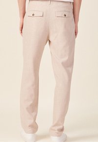 Pantalons en lin beige clair, coupe droite avec deux poches arrière et des boutons dorés, texture lisse, associés à des chaussures blanches.