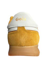 Las zapatillas Gola tienen una parte superior de cuero blanco y un talón de ante amarillo, con una suela de goma texturizada y un detalle del logo dorado en la pestaña del talón.