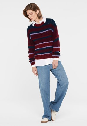Knit sweater with maroon, navy, and lilac stripes, round neckline, and dropped shoulders, worn over a white collared shirt and blue jeans.