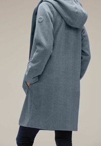 Ljusblå kappa med fiskbensmönster, huva, sidofickor och en cirkulär logotyp på överarmen. Mjuk textur och knälång design.