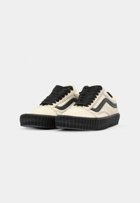 Zapatillas beige con acentos negros, con suelas de goma texturizada, punta redonda y cordones planos. Superficie lisa con rayas laterales en contraste.