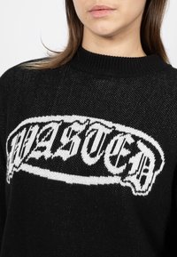 Frau trägt einen schwarzen Strickpullover mit weißem stilisiertem Text über der Brust, von den Schultern bis zur Mitte des Oberkörpers gezeigt.