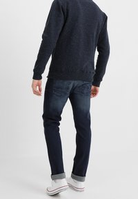 Maglione a maglia blu navy con motivo a macchie, abbinato a jeans aderenti blu scuro, con orlo risvoltato e sneakers bianche. Design minimalista con una texture sottile.