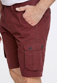 Burgundy cargoshorts med snedsträckta fickor fram, knappficka på sidan, platt midja och bomullstyg. Slät textur med subtil lyster.