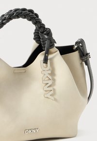 Cremefarvet DKNY håndtaske med et flettet sort læderhåndtag, sølvfarvede logo detaljer og en blød tekstureret overflade.