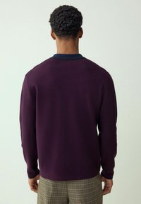 Rövid, göndör hajú férfi sötétlila hosszú ujjú ingben és bézs kockás nadrágban, háttal áll a kamerának.