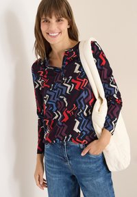 Lächelnde Frau in einem langärmligen Shirt mit einem bunt gestreiften Zickzack-Muster, blauen Jeans und einer weißen gepolsterten Schultertasche.
