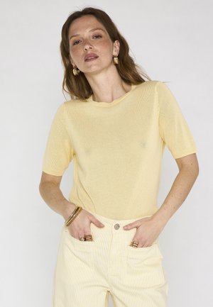 Donna con capelli castani lunghi che indossa orecchini a cerchio dorati, una maglietta gialla a maniche corte, pantaloni gialli a righe e braccialetti d'oro con le mani in tasca.