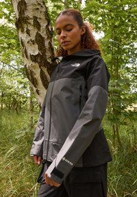 Gore-Tex jakna v črni in sivi barvi, z zadrgo spredaj, nastavljivo kapuco in ježkovimi manšetami. Lahka tkanina z gladko teksturo.