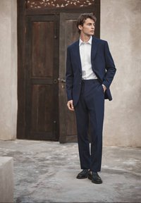 Jeune homme portant un costume bleu marine, une chemise blanche et des chaussures noires, debout avec une main dans la poche devant une porte en bois rustique.