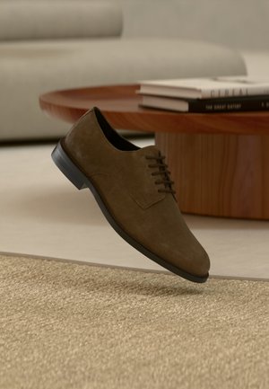 Καφέ σουέντ ανδρικό επίσημο παπούτσι με μαύρη σόλα που αιωρείται πάνω από μπεζ υφαντό χαλί, δίπλα σε ξύλινο τραπεζάκι καφέ με στοίβες βιβλίων.